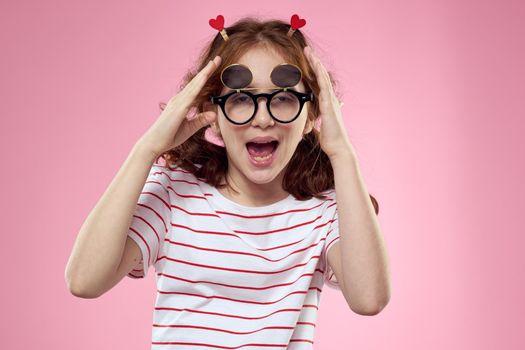 Cheerful little girl in fashionable glasses hairstyle decoration striped t-shirt pink background. High quality photo