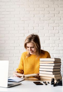 Young smiling woman in yellow sweater studying using laptop and reading a book