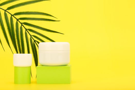 Face cream or mask and lip balm, scrub or under eye cream on green podiums against yellow background with palm leaf and copy space. Concept of sunscreen, summer skin care 