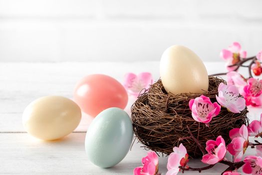 Close up of colorful Easter eggs in the nest with pink plum flower on bright white wooden table background.