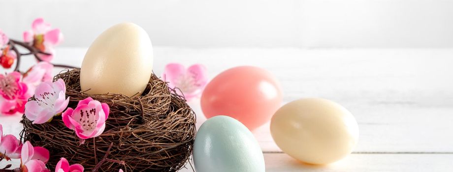 Close up of colorful Easter eggs in the nest with pink plum flower on bright white wooden table background.