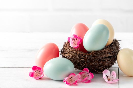 Close up of colorful Easter eggs in the nest with pink plum flower on bright white wooden table background.