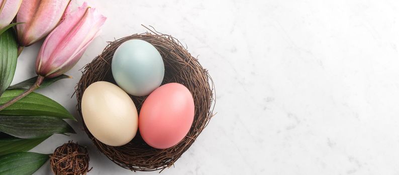 Colorful Easter eggs in the nest with pink lily flower on bright marble white table background.