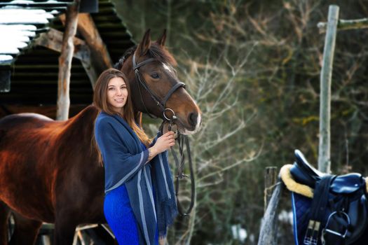 A beautiful girl in a blue dress and a stole stands next to a horse on the background of a forest and wooden buildings
