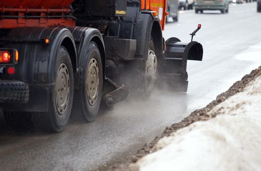 orange street cleaning truck with spinning brushes and dirt splashes at winter day - close-up with selective focus