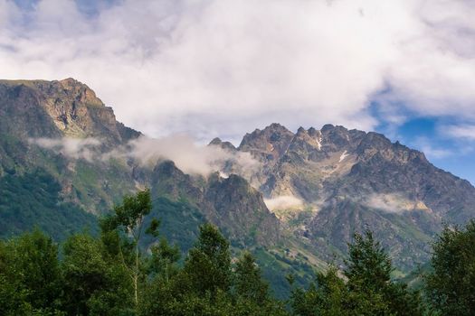 High mountains of the Caucasus with a beautiful view. Green vegetation and shrubs on sharp rocks above a blue sky. Magnificent mesmerizing landscape.