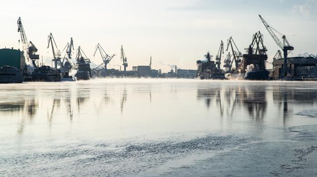 Cranes of of the Baltic shipyard on a frosty winter day, steam over the Neva river, smooth surface of the river, mirror reflection on the water, ships under construction, trawlers, nuclear icebreakers. High quality photo