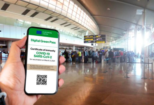 The digital green pass of the european union with the QR code on the screen of a mobile held by a hand with blurred airport in the background. Immunity from Covid-19. Travel without restrictions.