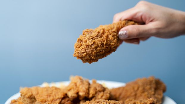 Hand holding drumsticks, crispy fried chicken in white plate on blue background.