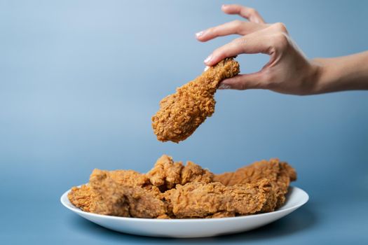 Hand holding drumsticks, crispy fried chicken in white plate on blue background.