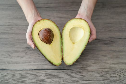 Woman hand holding cut half, sliced fresh green avocado on brown wooden table. Fruits healthy food concept.