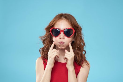 Little girl in sunglasses in the form of hearts red dress curly hair blue background. High quality photo