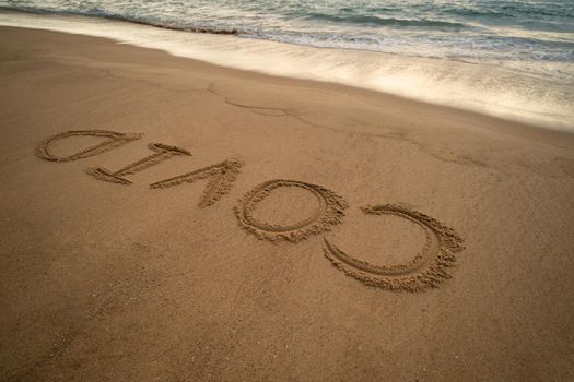 Handwriting COVID on sand and foam wave on beach. Coronavirus concept.