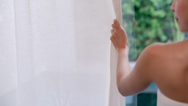 Woman hand opening white curtains window, green garden view outside.