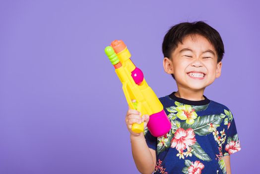 Happy Asian little boy holding plastic water gun, Thai kid funny hold toy water pistol and smiling, studio shot isolated on purple background, Thailand Songkran festival day national culture party