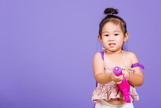 Thai child funny hold toy water pistol and smile, Happy Asian little girl holding plastic water gun, studio shot isolated on purple background, Thailand Songkran festival day national culture concept