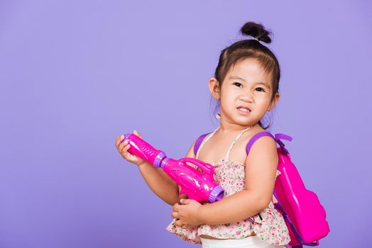 Thai child funny hold toy water pistol and smile, Happy Asian little girl holding plastic water gun, studio shot isolated on purple background, Thailand Songkran festival day national culture concept