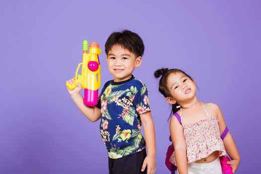 Two Happy Asian little boy and girl holding plastic water gun, Thai children funny hold toy water pistol and smile, studio shot isolated on purple background, Thailand Songkran festival day culture.