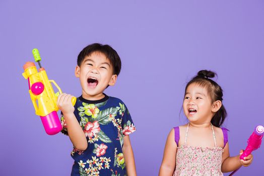 Two Happy Asian little boy and girl holding plastic water gun, Thai children funny hold toy water pistol and smile, studio shot isolated on purple background, Thailand Songkran festival day culture.