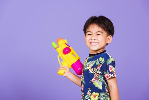 Thai kid funny hold toy water pistol and smiling, Happy Asian little boy holding plastic water gun, studio shot isolated on purple background, Thailand Songkran festival day national culture concept