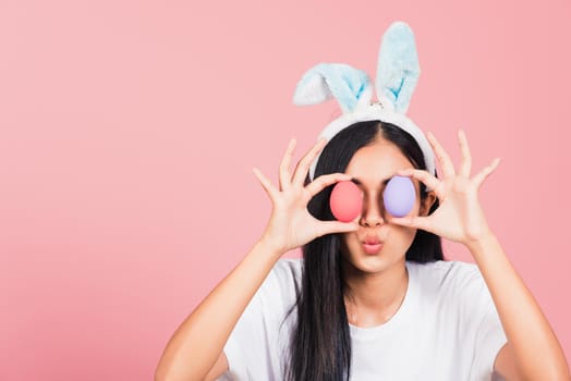 Happy beautiful young woman smiling wearing rabbit ears holding colorful Easter eggs front eyes, Thai female with bunny ear hold easter egg covering eye, studio shot isolated on pink background