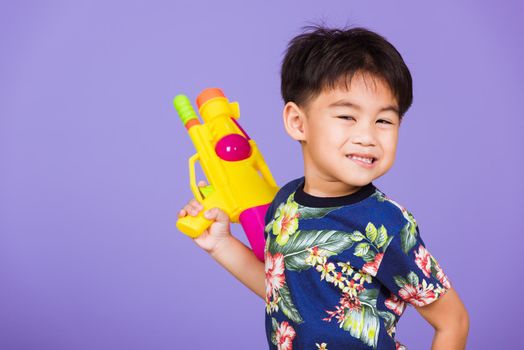 Thai kid funny hold toy water pistol and smiling, Happy Asian little boy holding plastic water gun, studio shot isolated on purple background, Thailand Songkran festival day national culture concept