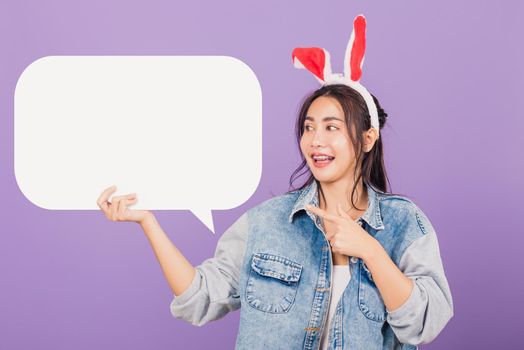 Happy Easter Day. Beautiful young woman smiling excited wearing rabbit ears and denims holding empty speech bubble, Portrait female looking at bubbles, studio shot isolated on purple background