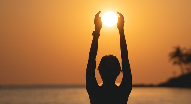 Sunset ocean view. Sun above the sea on orange sky. Woman standing on beach holding sun in her hands.