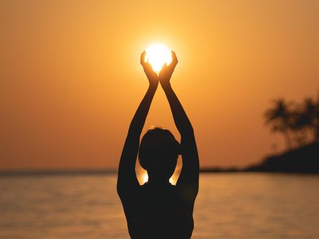 Sunset ocean view. Sun above the sea on orange sky. Woman standing on beach holding sun in her hands.