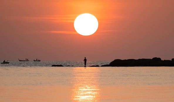 Sunset ocean view. Sun above the sea on orange sky. Fisherman with fishing rod on the rock.