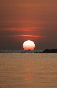 Sunset ocean view. Sun above the sea on orange sky. Fisherman with fishing rod on the rock.