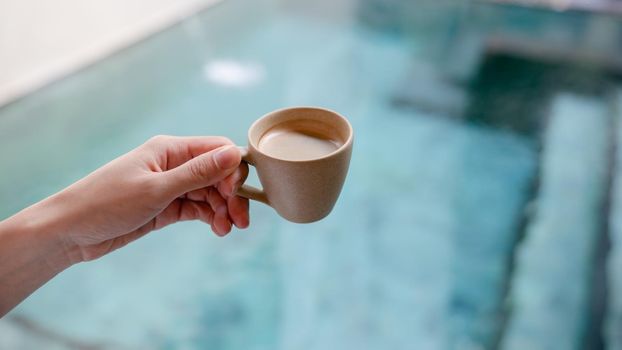 Woman hand holds a mug of hot coffee over the pool.