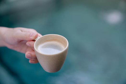 Woman hand holds a mug of hot coffee over the pool.