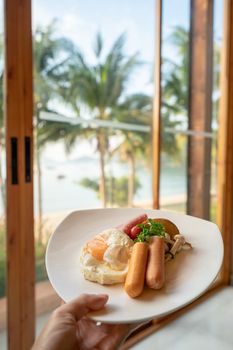 hand holding American breakfast set with fried eggs grill sausage and vegetable salad on beach background in morning.