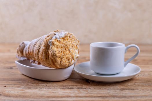 A cup of coffee next to a delicious aromatic puff pastry with protein cream, a delicious breakfast or snack on wooden table.