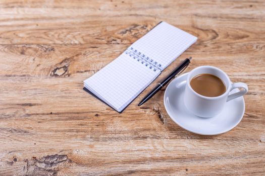 Small white coffee cup on a saucer stands on a wooden table next to a notebook and a pen, concept of coffee break at work, meeting, taking notes or planning
