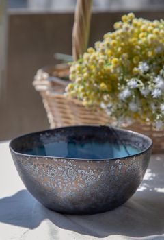 Ceramic bowl and bouquet of flowers in woven basket on white textured table cloth. Selective focus.