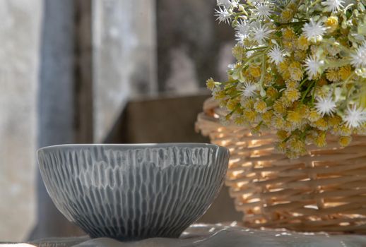 Ceramic bowl and bouquet of flowers in woven basket on white textured table cloth. Selective focus.