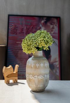 Bouquet of flowers in Beautiful ceramic vase and Wooden doll cat shape on white textured table cloth in front of Classic chinese poster movie frame with old cement wall. Home decor, Selective focus.