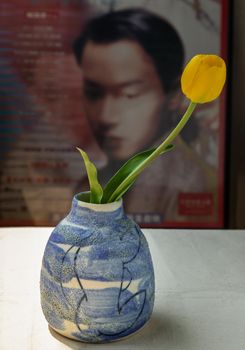 Bangkok, Thailand -Feb 06, 2021 : Yellow flower in Blue ceramic vase on white textured table cloth in front of Classic chinese poster movie frame. Home decor, Selective focus.