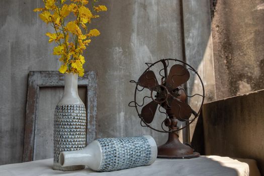 Yellow bouquet of flowers in White ceramic vase bottle shape and Vintage brown metal fan on white textured table cloth with old cement wall at the balcony house. Home decor, Selective focus.