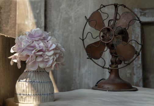 Bouquet of Purple flowers in White ceramic vase and Vintage brown metal fan on white textured table cloth with old cement wall at the balcony house. Home decor, Selective focus.