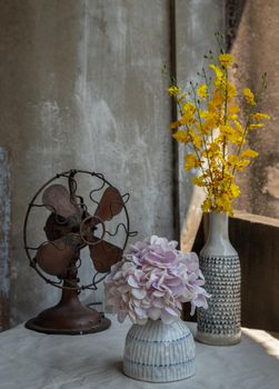 Bouquet of flowers in Two ceramic vase and Vintage brown metal fan on white textured table cloth with old cement wall at the balcony house. Home decor, Selective focus.