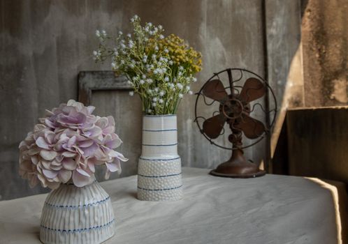 Bouquet of flowers in Two white handmade ceramic vase and Vintage brown metal fan on white textured table cloth with old cement wall at the balcony house. Home decor, Selective focus.