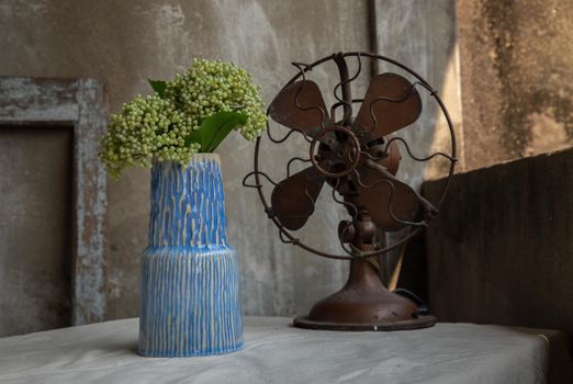 Bouquet of green flowers in Blue ceramic vase and Vintage brown metal fan on white textured table cloth with old cement wall at the balcony house. Home decor, Selective focus.