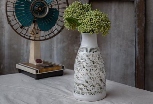 Bouquet of green flowers in White handmade ceramic vase bottle shape and Vintage fan on white textured table cloth with old cement wall. Home decor, Selective focus.