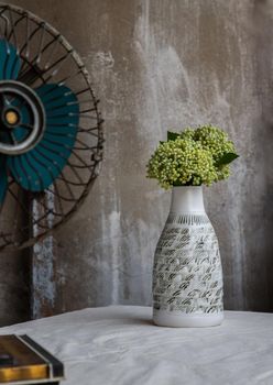 Bouquet of green flowers in White handmade ceramic vase bottle shape and Vintage fan on white textured table cloth with old cement wall. Home decor, Selective focus.