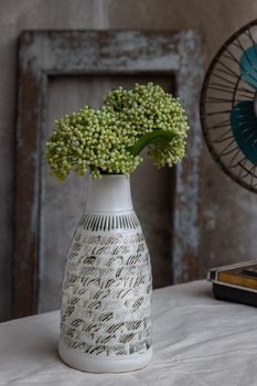 Bouquet of green flowers in White handmade ceramic vase bottle shape and Vintage fan on white textured table cloth with old cement wall. Home decor, Selective focus.