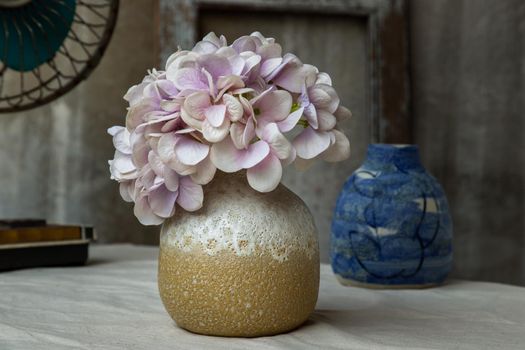 Bouquet of purple flowers in White with yellow handmade ceramic vase and Vintage fan on white textured table cloth with old cement wall. Home decor, Selective focus.