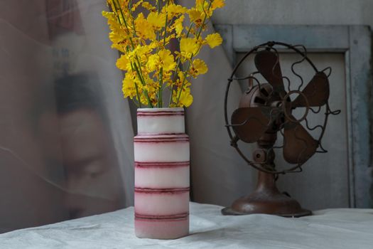 Bouquet of yellow flowers in Pink handmade ceramic vase and Vintage fan on white textured table cloth with old cement wall. Home decor, Selective focus.
